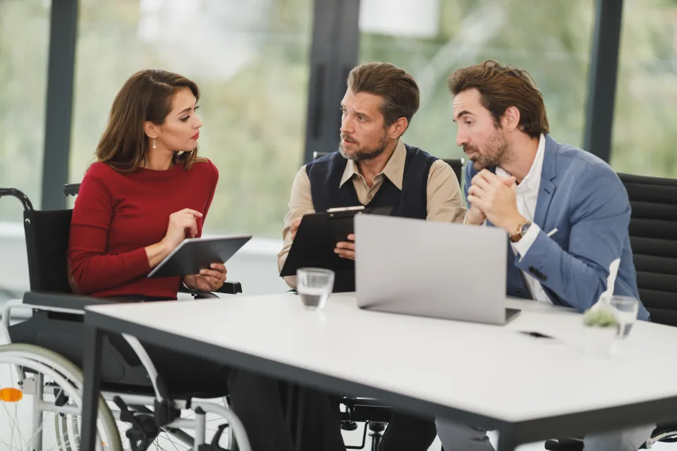Profissional em cadeira de rodas conversando com dois colegas em reunião, com tablet e notebook na mesa