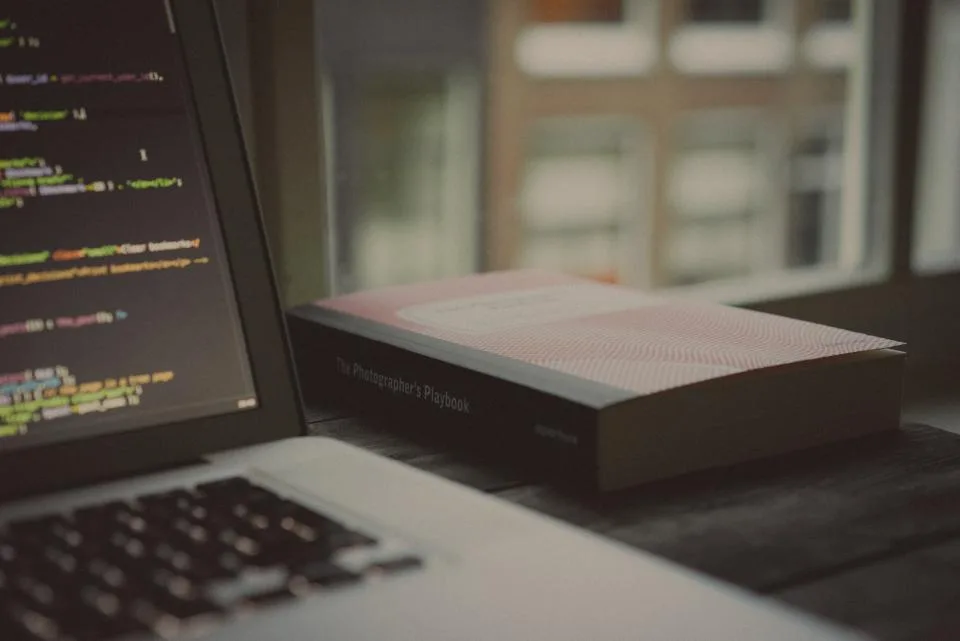 Notebook com código na tela ao lado de um livro, sobre uma mesa junto à janela