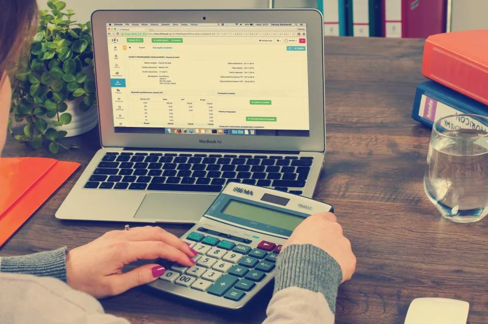 Mãos em uma calculadora ao lado de um notebook com planilha de cadastro aberta sobre a mesa