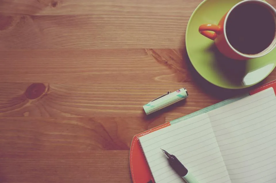 Caderno aberto com anotações, caneta e xícara de café sobre uma mesa de madeira