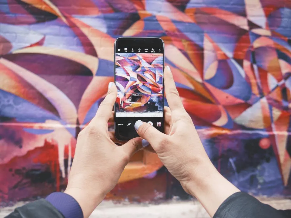 Mãos segurando um celular que fotografa um mural colorido
