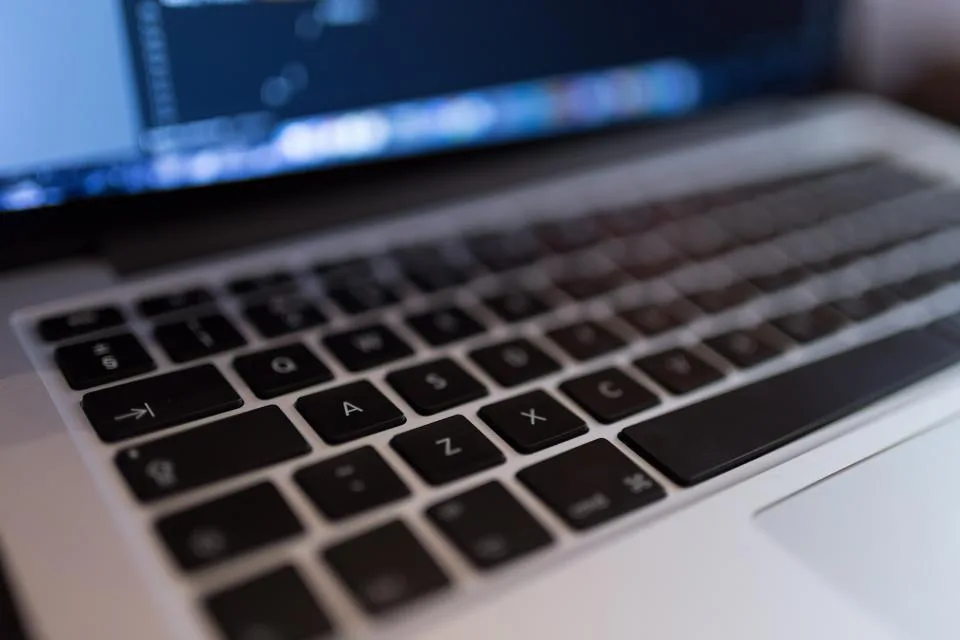 Teclado de notebook em close com código na tela ao fundo