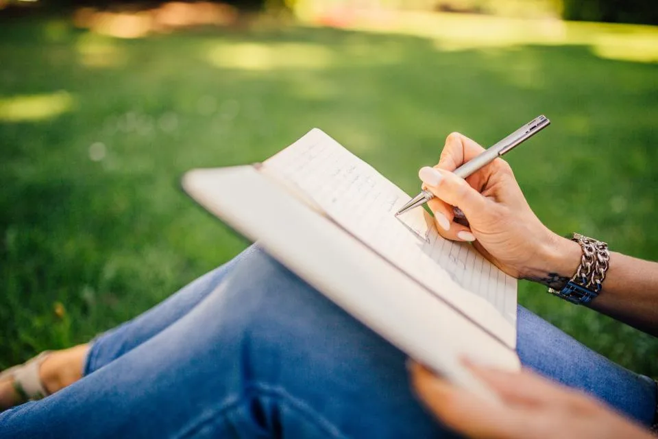 Pessoa escrevendo à mão num caderno, sentada na grama ao ar livre