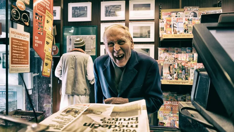 Jornaleiro sorrindo numa banca, com jornais expostos sobre o balcão