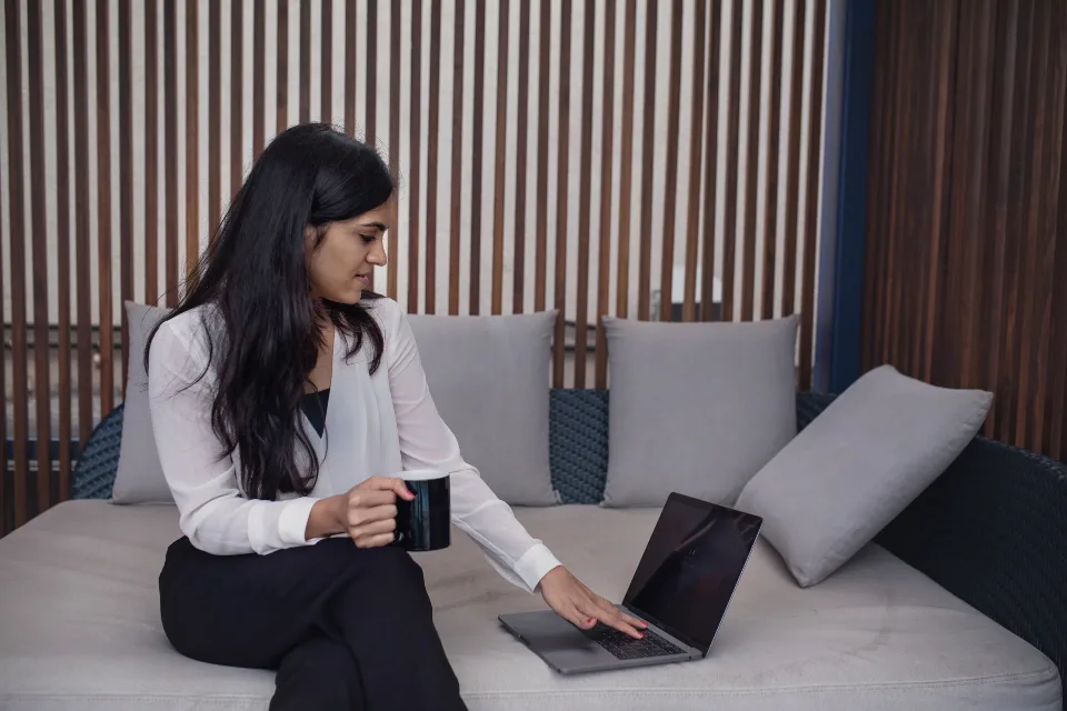 Mulher com uma caneca de café usando o notebook, sentada num sofá