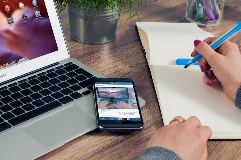Pessoa escrevendo à mão num caderno ao lado de um notebook e de um celular