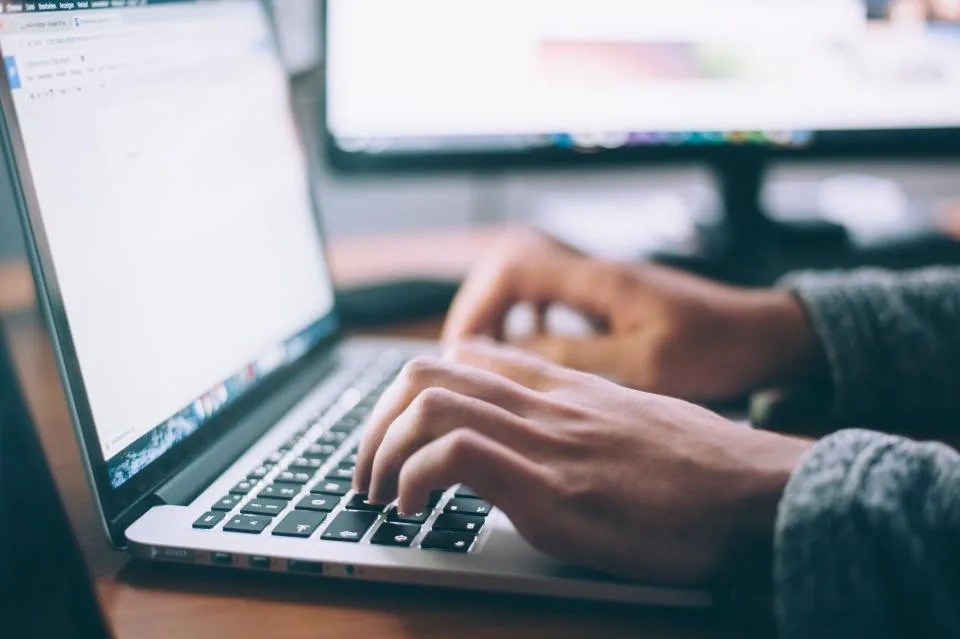 Mãos digitando no teclado de um notebook com um texto aberto na tela