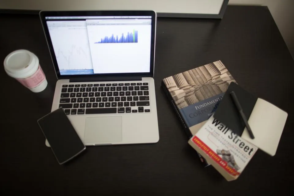 Mesa escura com notebook exibindo um gráfico de barras, celular, caderno e livros de finanças ao lado de um copo de café