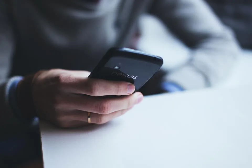 Mão segurando um smartphone apoiado na mesa, em primeiro plano