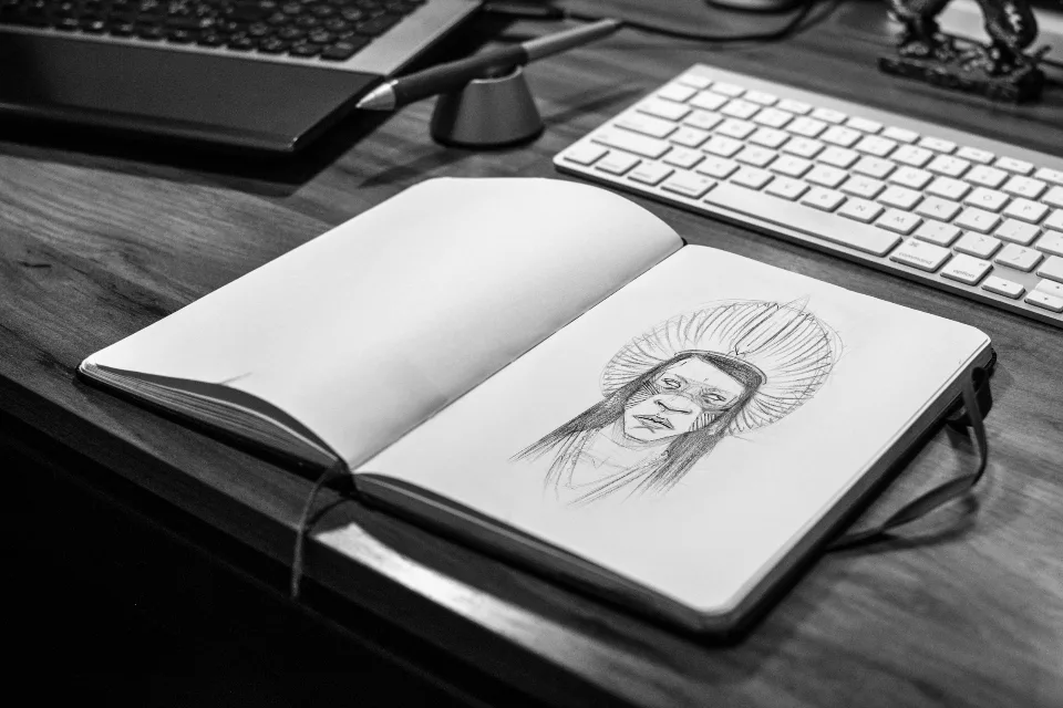 Caderno de desenho aberto sobre a mesa, com um esboço a lápis, ao lado de um teclado e de uma caneta digital