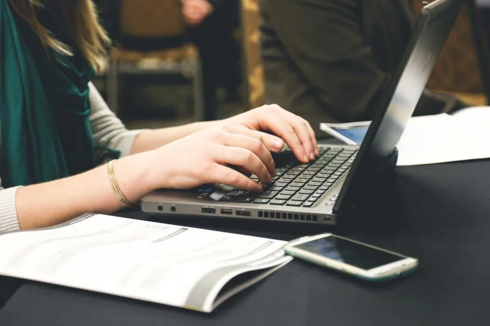 Mãos digitando num notebook, com um celular e papéis impressos sobre a mesa