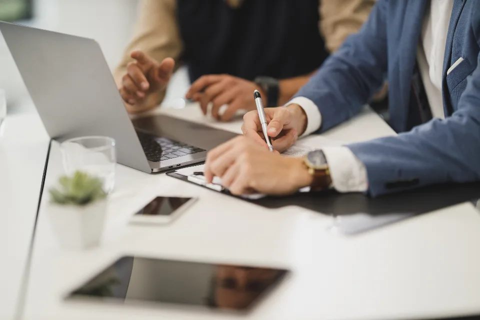 Duas pessoas em reunião à mesa, uma anotando respostas num caderno ao lado do notebook