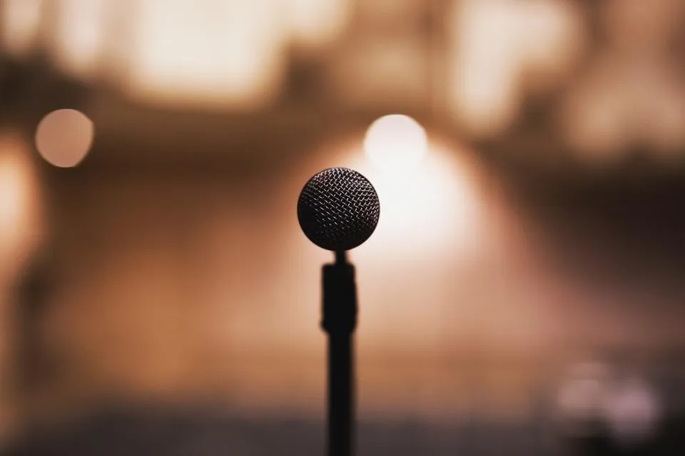 Microfone em pedestal diante de um palco com luzes desfocadas, à espera de quem vai falar