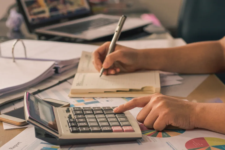 Mãos usando calculadora sobre relatórios com gráficos de pizza e uma pessoa anotando num caderno